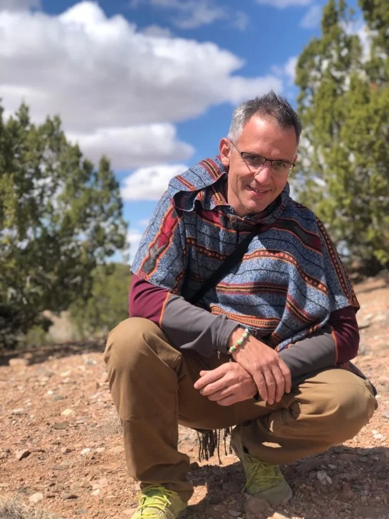 Carlos del Río en cuclillas sobre tierra seca, con poncho andino, entre árboles